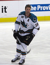 Hockey player in white and blue uniform without his helmet on. He skates across the ice, holding his stick off the ground.