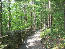A gravel path through a mixed forest of deciduous and conifer trees, with a rail fence supported by stone pillars left of the path