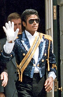 A mid-twenties African American man wearing a sequined military jacket and dark sunglasses. He is walking while waving his right hand, which is adorned with a white glove. His left hand is bare.