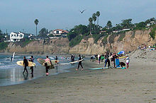 Several people, some wearing full length suits and carrying surf boards, on a beachfront with houses visible above them