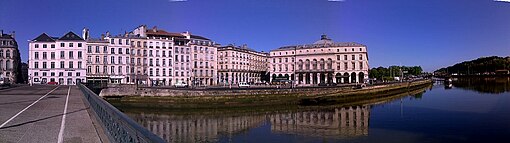 Vue panoramique de bâtiments à façade blanche se reflétant dans un cours d’eau sous un ciel bleu intense.