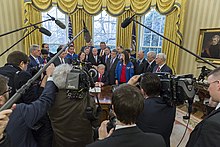 Trump, seated at the Resolute Desk in the White House, speaking to a crowd of reporters with boom microphones in front of him and public officials behind him