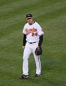 A man in a white baseball uniform with orange script across the chest and a black baseball cap walks across a grass field with a black baseball glove on his left hand