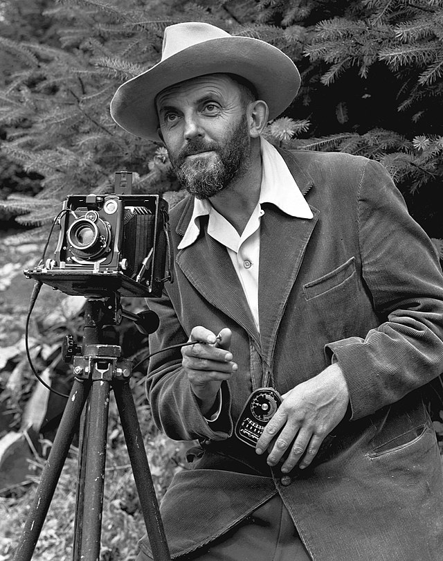 A photo of a bearded Ansel Adams with a camera on a tripod and a light meter in his hand.  Adams is wearing a dark jacket and a white shirt, and the open shirt collar is spread over the lapel of his jacket.  He is holding a cable release for the camera, and there is a rocky hillside behind him.  The photo was taken by J. Malcolm Greany, probably in 1947.