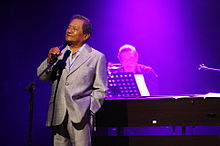 A man facing left is performing on a stage with a microphone in his right hand.