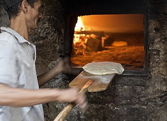 Мужчина с помощью кожуры хлеба опускает круглый диск сырого теста для лепешек в кирпичную печь.