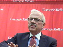 Barr, visible from the chest up, wears a navy blue jacket with blue shirt and red and blue checkered tie, in front of a red background with the words "Carnegie Mellon" written on it in several places