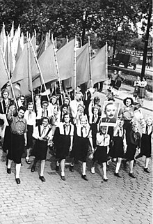 Photo noir et blanc d'un groupe de jeunes filles en chemise blanche et juppe noire, défilant dans une rue tout en agitant des drapeaux et le portrait d'un homme.