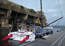 Voilier bleu sombre et blanc, vu de trois quarts arrière, amarré au ponton. En arrière-plan, l'ancienne base de sous-marins allemande de Lorient.