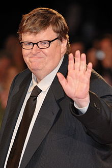 A mean wearing glasses and a suit waves to the camera and smiles slightly.