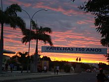 A street in Parelhas, wi palm trees an biggins, an a banner statin "150 Anos" (150 years) athort the street, at sunset