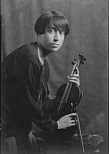 A young woman with short dark hair, leaning forward, holding a violin.