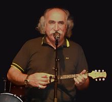 Un chanteur assez âgé, portant un t-shirt marron, chante devant un micro tout en jouant avec une sorte de petite guitare, appelé baglamas.