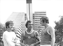 Photo en noir et blanc de trois athlètes est-Allemands souriants. En fond, on voit de profil une des demi-pyramides.