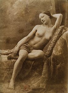 Braune Sepia-Fotografie von einem weiblichen Akt, die auf einer Chaiselongue sitzt. Mit ihrem linken Arm hält sie ihr Haar hinter dem Kopf zusammen und hat ihr linkes Bein unter ihren rechten Oberschenkel positioniert. Ein Tuch bedeckt ihren Intimbereich.
