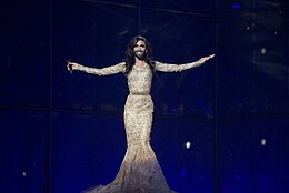 Een man met lang haar en een baard draagt een glitterende gouden jurk op een podium