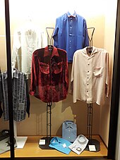 Plusieurs chemises colorées sont exposées dans une vitrine du musée, sur des présentoirs en métal dotés de cintres. Elles sont de couleurs variées : noire rayée de blanc, blanche, rouge façon velours dévoré, bleu marine, rose, turquoise, gris perle, blanc cassé.