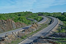 A divided four-lane highway curves between large granit roack outcroppings, passing below high-voltage powerlines in the background