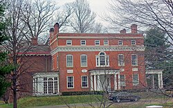 A large two-story flat-roofed rectangular brick building with four corner chimneys classically inspired ornamentation. There is a large full-height wing on the left and a small white wooden porch on the right. There are bare trees and a car parked in the front driveway.