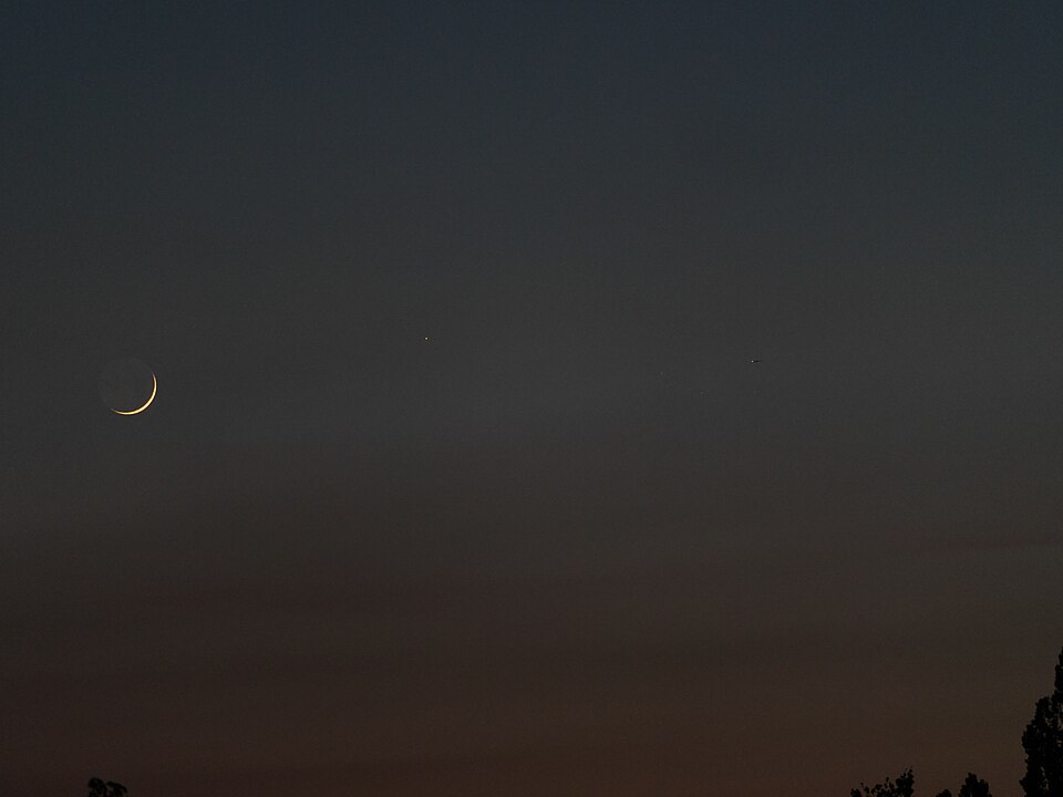 Vier Objekte bei einer Höhe von ungefähr 9 Bogengrad (von links nach rechts): Mond (Elongation = 21,4 Bogengrad, 3,5 Prozent beleuchtet), rechts davon Merkur (Elongation = 20,1 Bogengrad, 28 Prozent beleuchtet), weiter rechts die Plejaden (Höhe = 9 Bogengrad), direkt darüber ein Flugzeug. Die Sonne stand bereits über 8 Bogengrad unterhalb der Horizontlinie