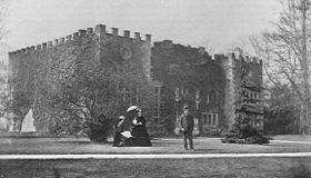 An old black-and white photograph showing a three-storey battlemented house, in front of which is a standing male and two sitting females