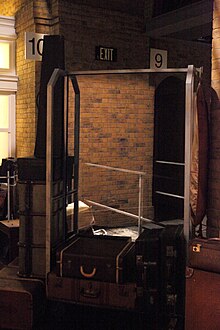 Photograph of a hotel-style metal-framed luggage trolley surrounded by old-style suitcases. In the background is what appears to be a solid brick wall. with platform number sign "10" on the left, and "9" on the right. The trolley contains a human-sized semi-transparent mirror that combines the foreground and background.