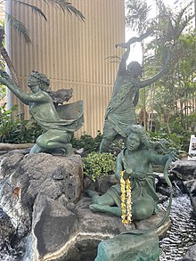Three bronze sculptures of women standing and sitting on rocks in a water fountain. The three figures hold cloth and beating sticks.