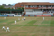  A ground with sportsman playing cricket, with stands in the background