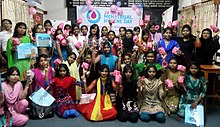 Gruppenbild vor einem Banner des Menstrual Hygiene Day, zum Großteil Frauen in traditioneller, bunter Bekleidung. Einige halten rosafarbene Blumen, Päckchen oder Luftballons in die Höhe.