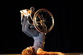 A woman in dark, close-fitting clothes and a wheelchair lies on her back, stabilizing herself with her arms and lifting her legs, along with the wheelchair, over her head