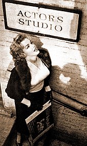 Monroe, who is wearing a skirt, blouse and jacket, standing below a sign for the Actors Studio looking up towards it