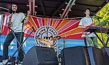 three young men on stage, one singing into a handheld microphone, one playing drums, and the other playing an electric guitar behind a keyboard