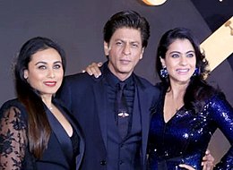 Shah Rukh Khan merangkul Rani Mukerji dan Kajol, dan mereka semua berdiri di atas sebuah karpet merah.