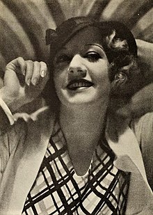 Ruth Selwyn, a smiling white woman with short blonde wavy hair, wearing a hat and a blouse with a graphic print