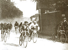Photographie en noir et blanc d'un groupe de cyclistes, l'un vêtu de blanc au centre.
