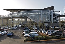 A tall and wide glass building with an angled roof, overlooking a parking lot full of cars.