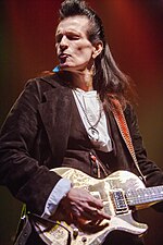 The head and torso of a middle-aged man. He has long, dark hair. He is wearing a dark jacket, a light top, and several items of jewelry, including large earrings. He is playing an electric guitar.