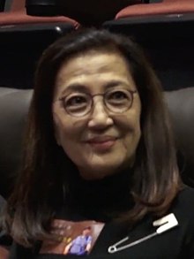 Ma, wearing a black shirt and glasses, facing right while seated in a cinema.