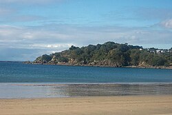 Fotografía de una playa con una pequeña colina en el fondo, ubicada en la Isla Waiheke en Nueva Zelanda. El pie de imagen dice: "Según el canal de noticias Newshub y la administración de la Isla Waiheke (en la imagen), una de las playas en esta isla fue utilizada como lugar de grabación del vídeo musical".