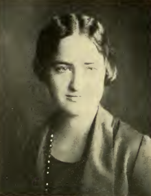 A young white woman with hair parted center, wearing a jacket and a strand of beads