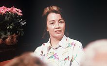 Фотография женщины с каштановыми волосами до плеч, обращенной к зрителю и смотрящей немного вправо, в белой рубашке с цветочным принтом.