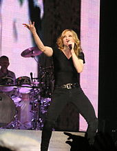 Блондинка-исполнительница поет в микрофон, поднимая левую руку.