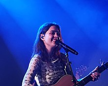 In blue lighting especially visible on her hair, a woman performs onstage with an acoustic guitar and a big smile.
