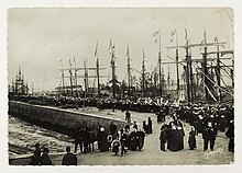 Vue du port de Paimpol un jour de pardon : à droite de la jetée, les goélettes sont décorées de fanion. Au bout de la jetée, la procession est réunie devant le reposoir supportant une statue de la Vierge Marie.