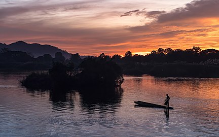 Silhouette d'un pêcheur debout sur sa pirogue au coucher du soleil avec nuages orange, à Don Det. Décembre 2020.