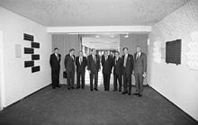 Eight men in suits stand in a hall facing forward.