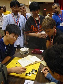 Three Japanese professional Go players observe some younger amateurs as they dissect a life and death problem in the corner of the board, at the US Go Congress in Houston, Texas, 2003. Go pros and amateurs.jpg.jpeg