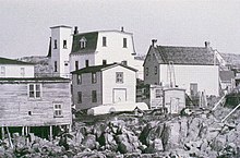 The photo shows a rocky foreground, then three clapboarded houses with assorted outbuildings. The Greenspond Courthouse, a large building with a central tower and mansard roof, is in the background.
