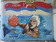 Auf dem bemalten Mauerabschnitt hält Nikić den Ball in seiner rechten Hand. Er trägt eine weiße Badekappe mit der Nummer 7 und der Aufschrift SRB für Serbien.