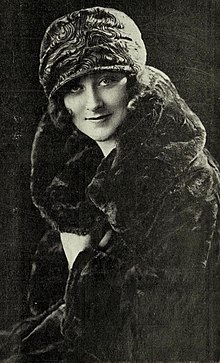 A young white woman wearing a dark fur coat and a printed fabric hat, worn low over her brow.
